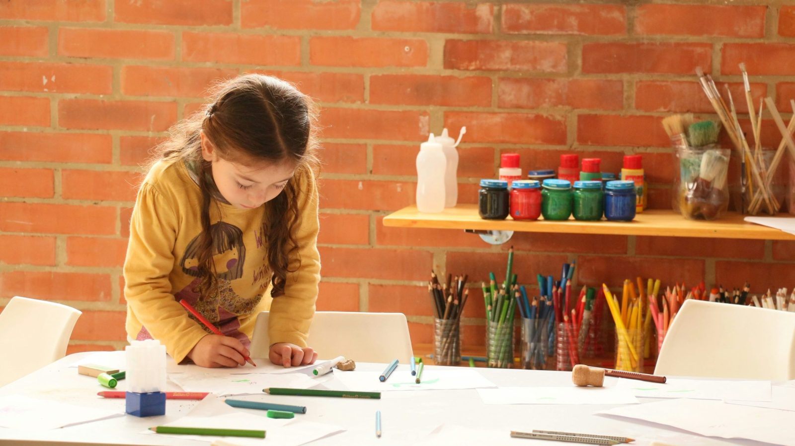 enfant qui dessine sur une table avec en fond un set de pinceaux, crayons et peinture